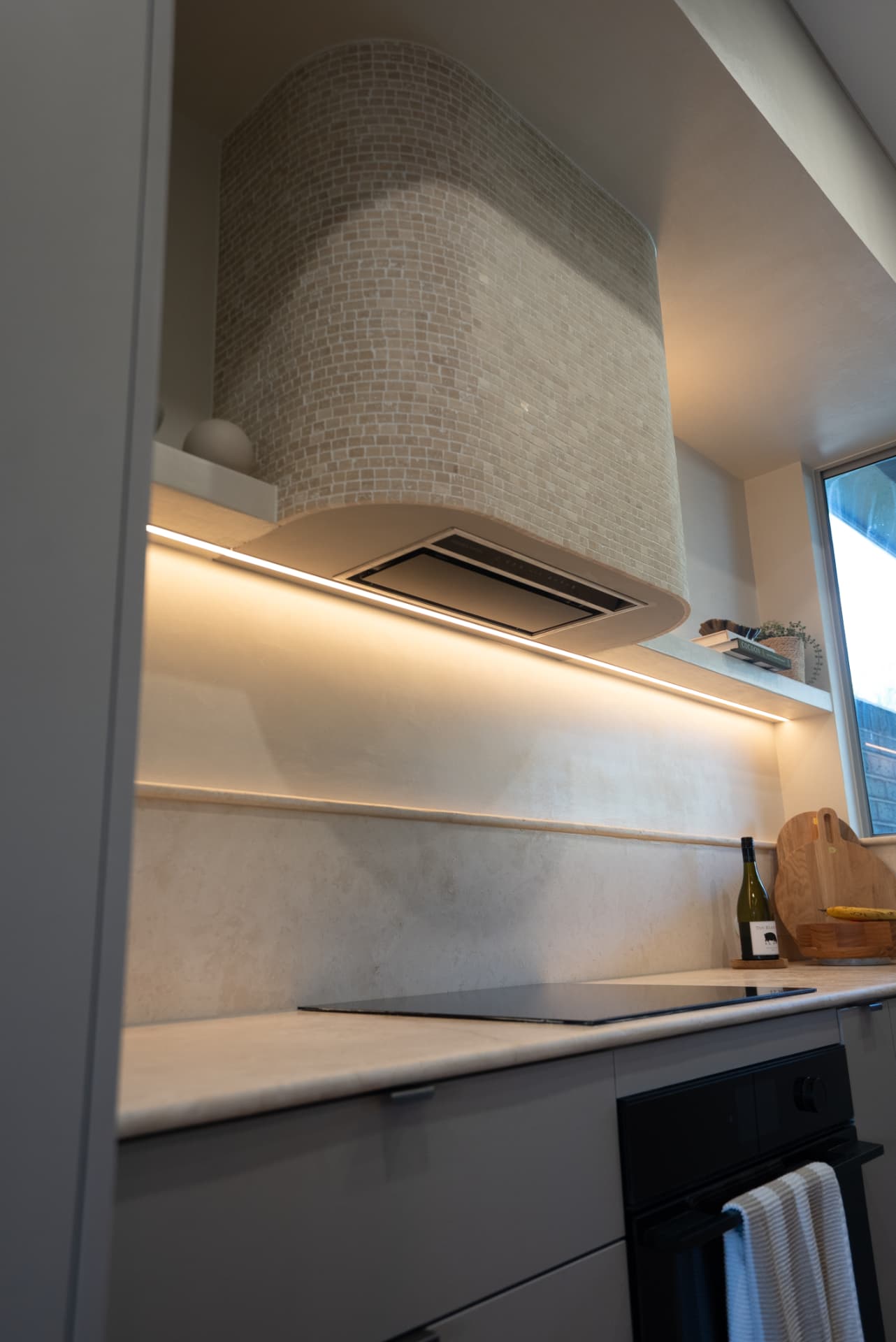 Curved tiled rangehood detail — Mosaic-tiled curved hood with recessed LED strip lighting over the cooktop.